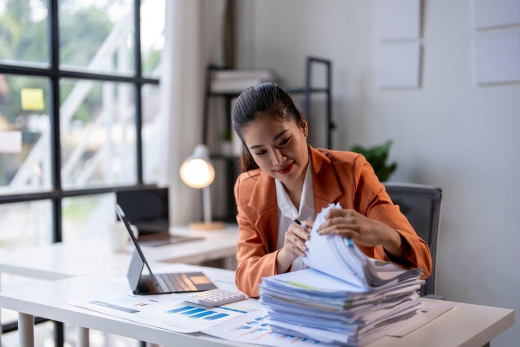 Businesswoman searching for documents in stack of paperwork at office desk